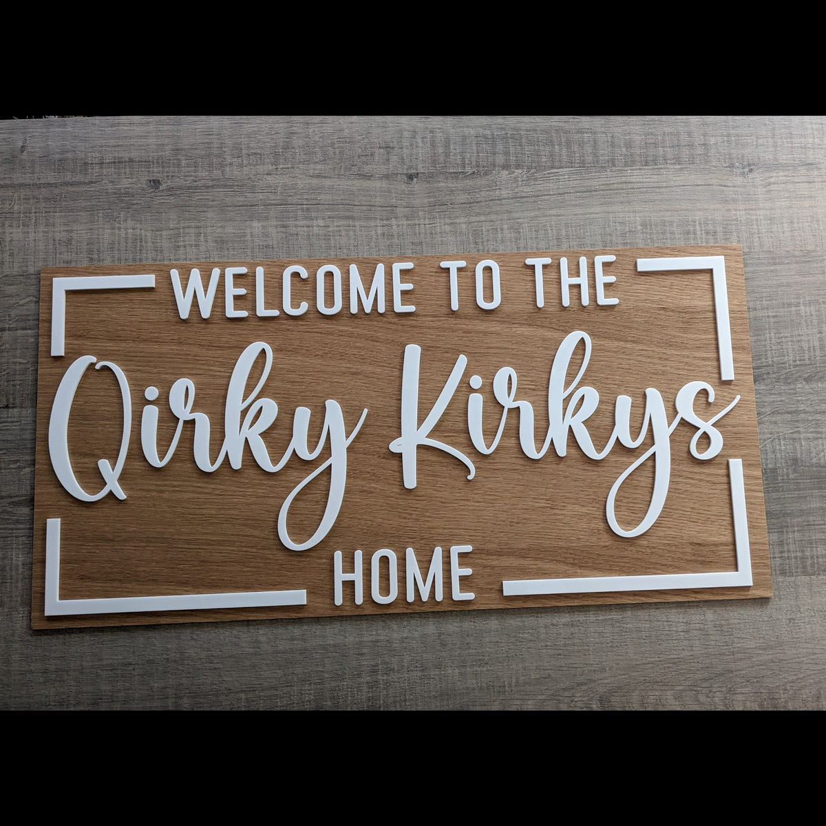 A rectangular wooden sign with white lettering that reads 'WELCOME TO THE Qirky Kirkys HOME' and white corner brackets, displayed on a gray wood-textured surface.