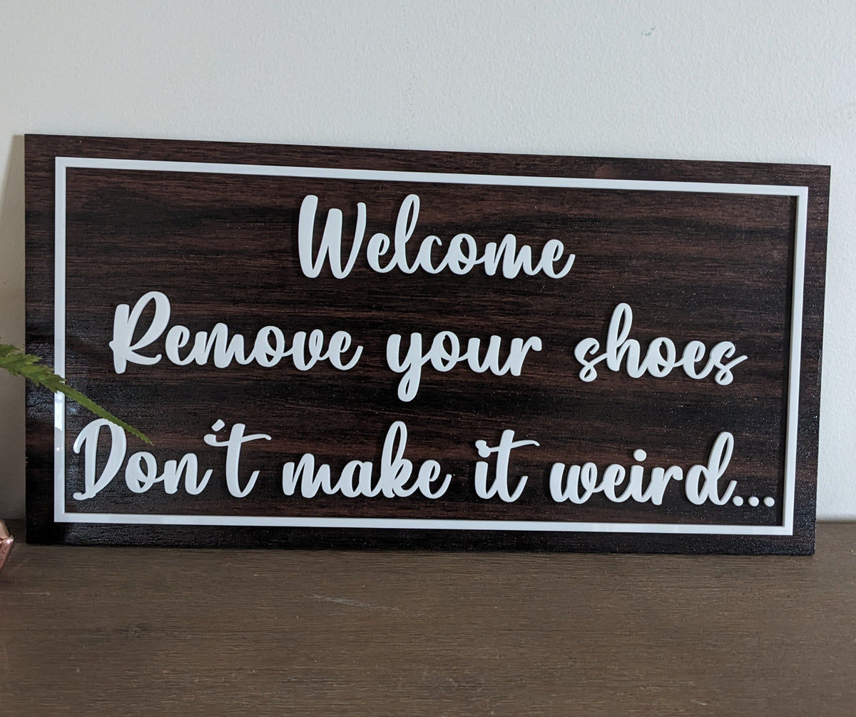 A rectangular dark wood sign with white lettering that reads 'Welcome Remove your shoes Don't make it weird.