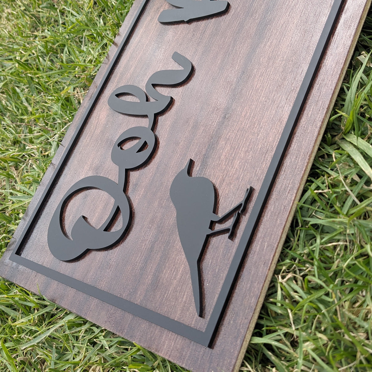 A rectangular wooden sign with a dark brown wood grain finish, featuring a black metal border and custom cutout lettering and silhouette designs, shown lying on green grass.