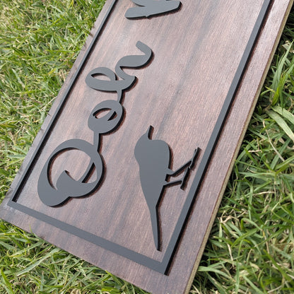 A rectangular wooden sign with a dark brown wood grain finish, featuring a black metal border and custom cutout lettering and silhouette designs, shown lying on green grass.