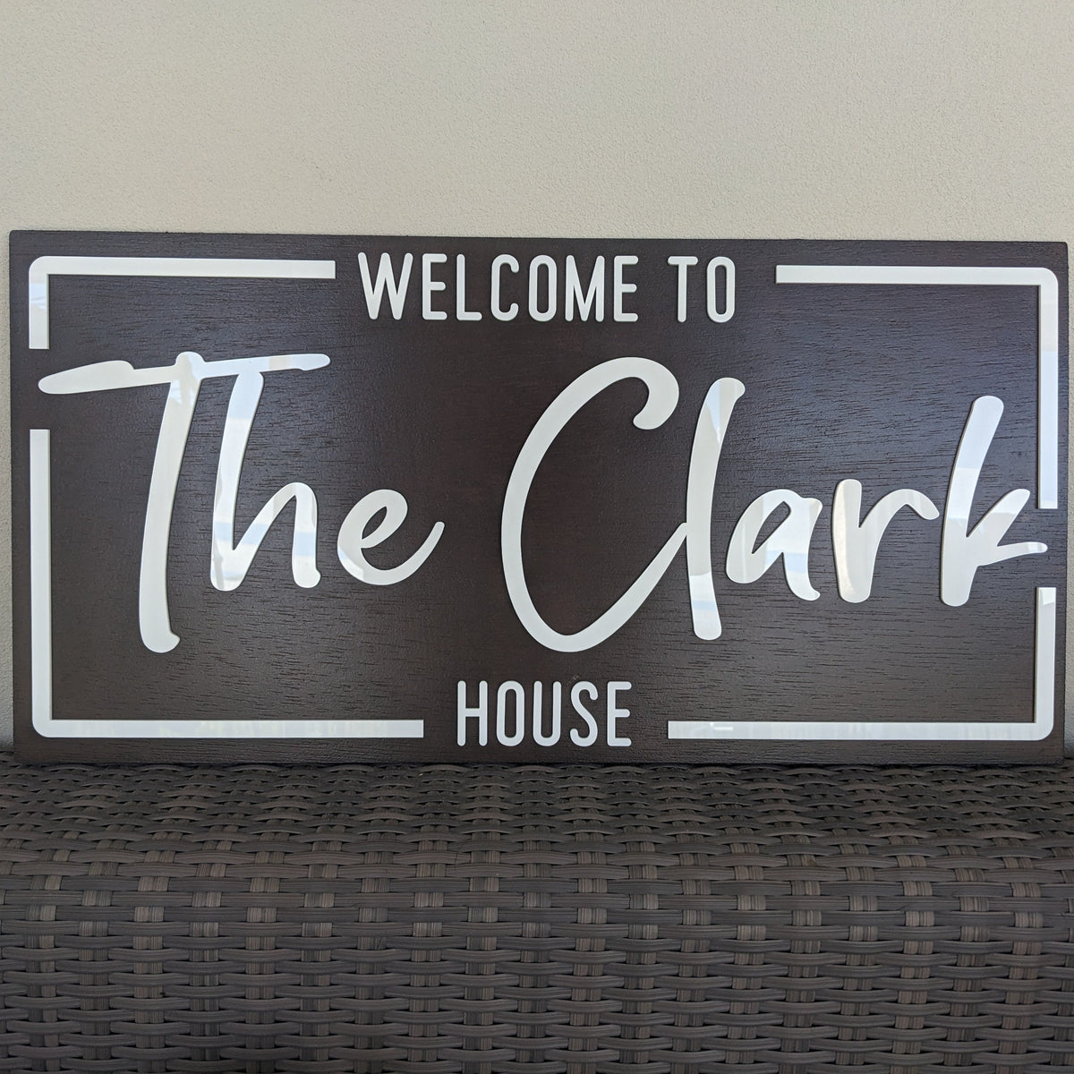 A rectangular dark brown wooden sign with white lettering that reads 'WELCOME TO The Clark HOUSE', displayed against a light beige wall above a woven dark surface.