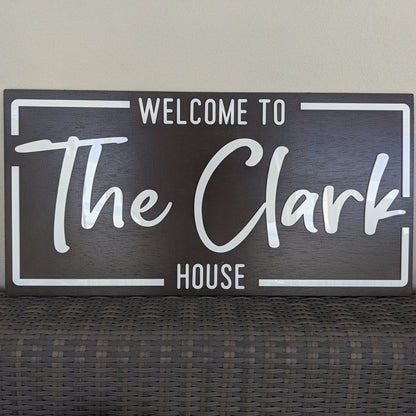 A rectangular dark brown wooden sign with white lettering that reads 'WELCOME TO The Clark HOUSE', displayed against a light beige wall above a woven dark surface.