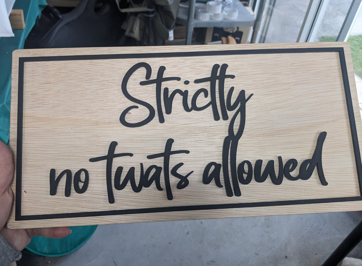 A hand holds a rectangular wooden sign with the phrase 'Strictly no twats allowed' in black script and border, photographed in a real indoor setting with a table and window in the background.
