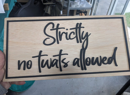 A hand holds a rectangular wooden sign with the phrase 'Strictly no twats allowed' in black script and border, photographed in a real indoor setting with a table and window in the background.