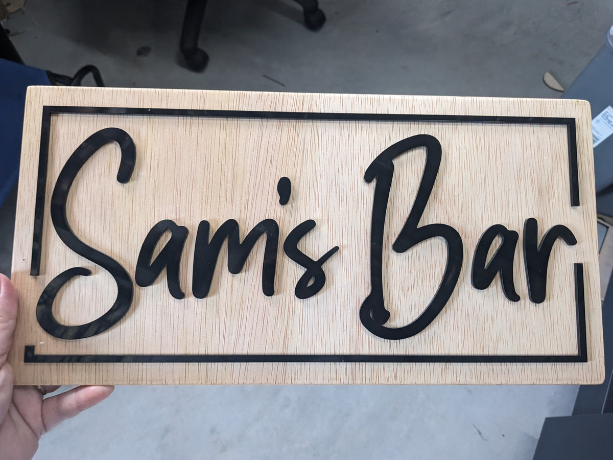A rectangular wooden sign with the words 'Sam's Bar' in black cursive lettering, held by a hand against a concrete floor background.