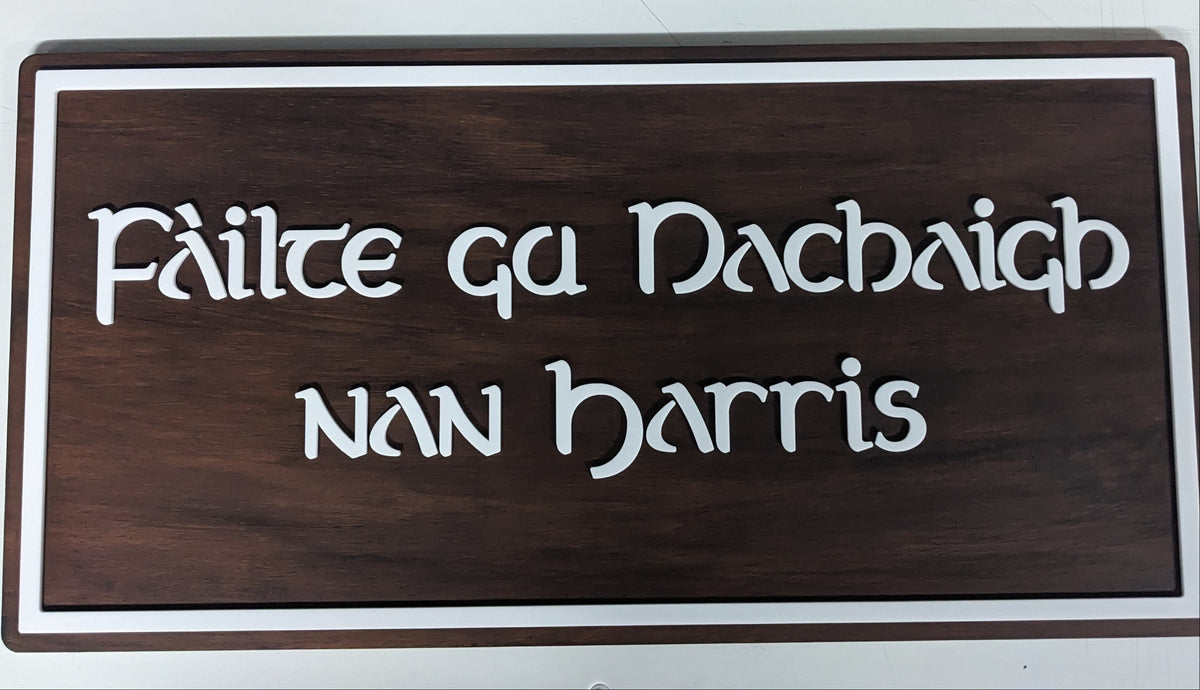 A rectangular wooden sign with a dark brown wood grain finish, featuring the Gaelic phrase 'Fáilte gu Nachaigh nan Harris' in raised white lettering, bordered by a white frame.