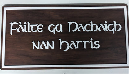 A rectangular wooden sign with a dark brown wood grain finish, featuring the Gaelic phrase 'Fáilte gu Nachaigh nan Harris' in raised white lettering, bordered by a white frame.
