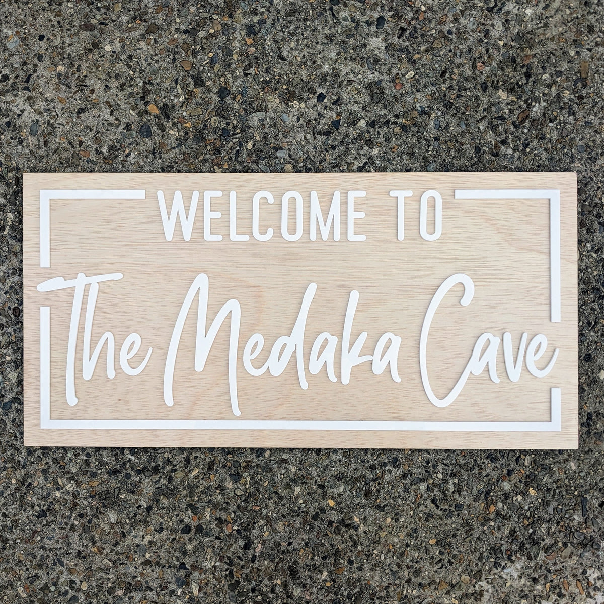 A rectangular light wood sign with white lettering that reads 'WELCOME TO The Medaka Cave,' placed on a speckled gray stone surface.