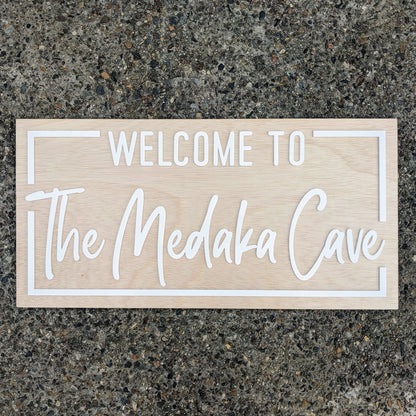 A rectangular light wood sign with white lettering that reads 'WELCOME TO The Medaka Cave,' placed on a speckled gray stone surface.