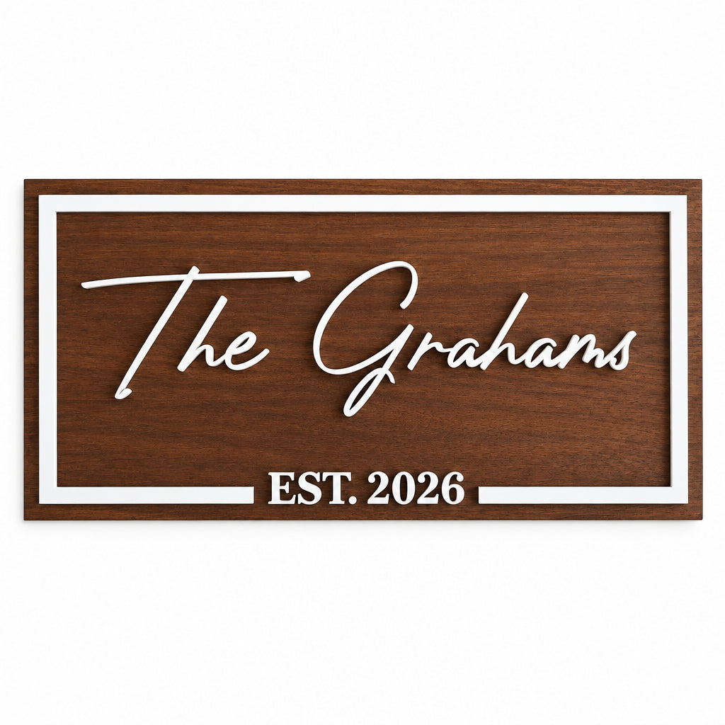 A personalized wooden sign with a dark brown wood grain finish, featuring white raised lettering that reads 'The Grahams' and 'EST. 2026', bordered by a white frame, shown mounted on a white wall.