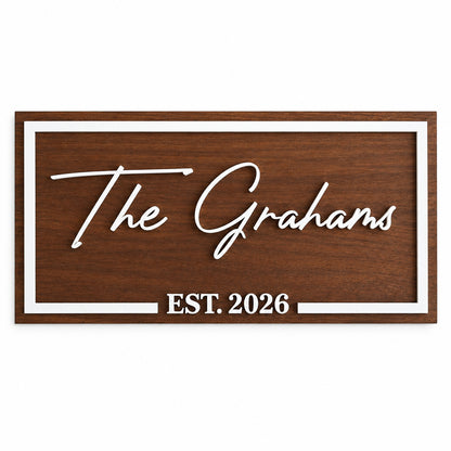 A personalized wooden sign with a dark brown wood grain finish, featuring white raised lettering that reads 'The Grahams' and 'EST. 2026', bordered by a white frame, shown mounted on a white wall.