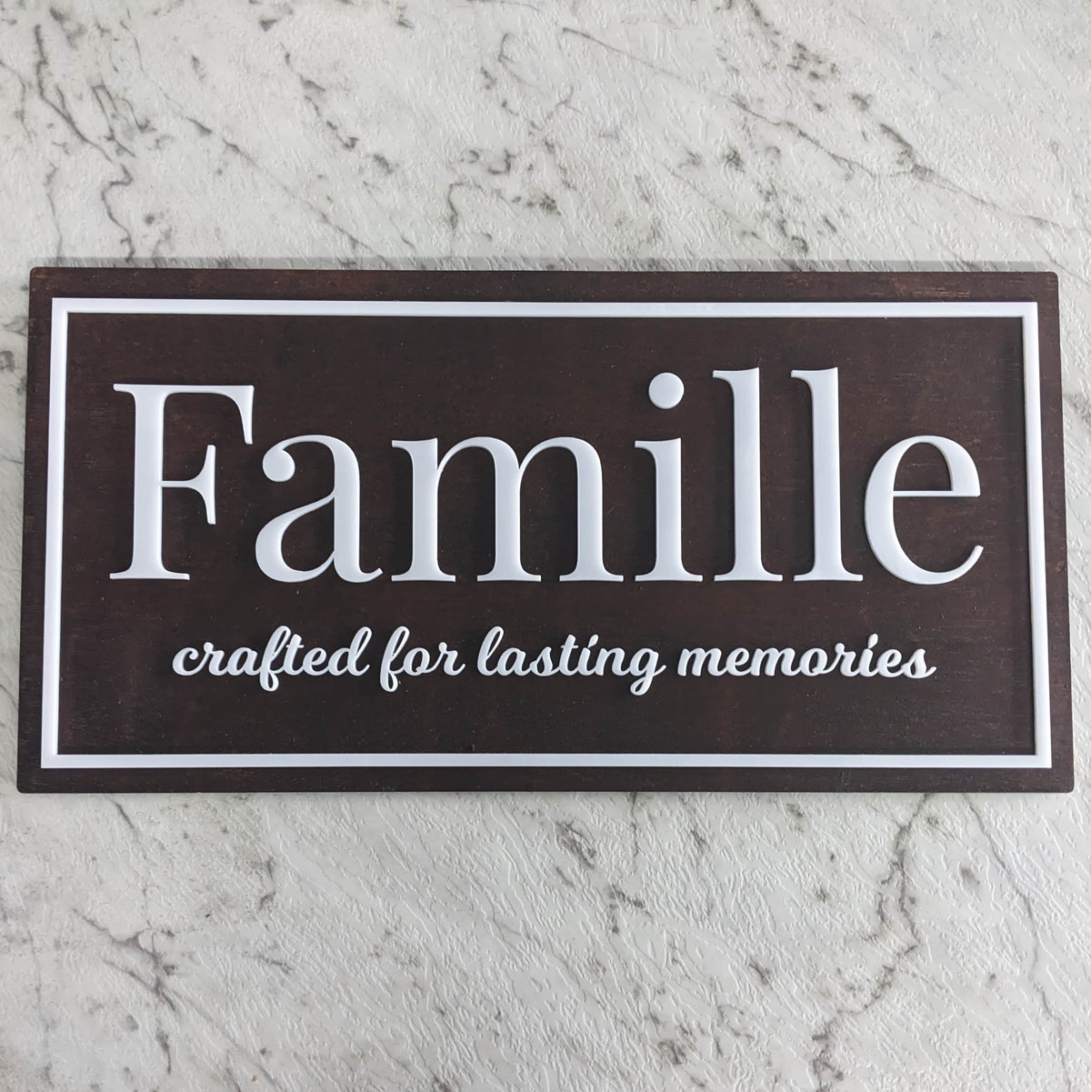A rectangular dark brown wooden sign with white text reading 'Famille' and 'crafted for lasting memories', displayed on a light gray marble surface.