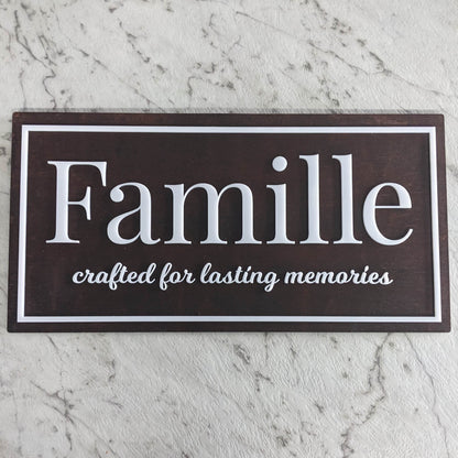A rectangular dark brown wooden sign with white text reading 'Famille' and 'crafted for lasting memories', displayed on a light gray marble surface.