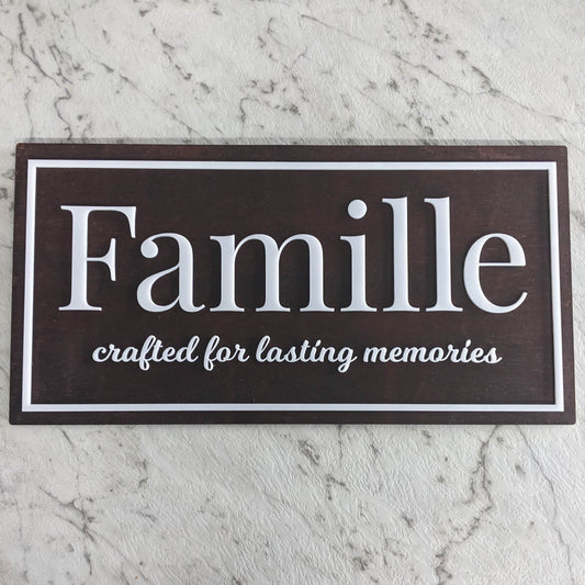 A rectangular dark brown wooden sign with white text reading 'Famille' and 'crafted for lasting memories', displayed on a light gray marble surface.
