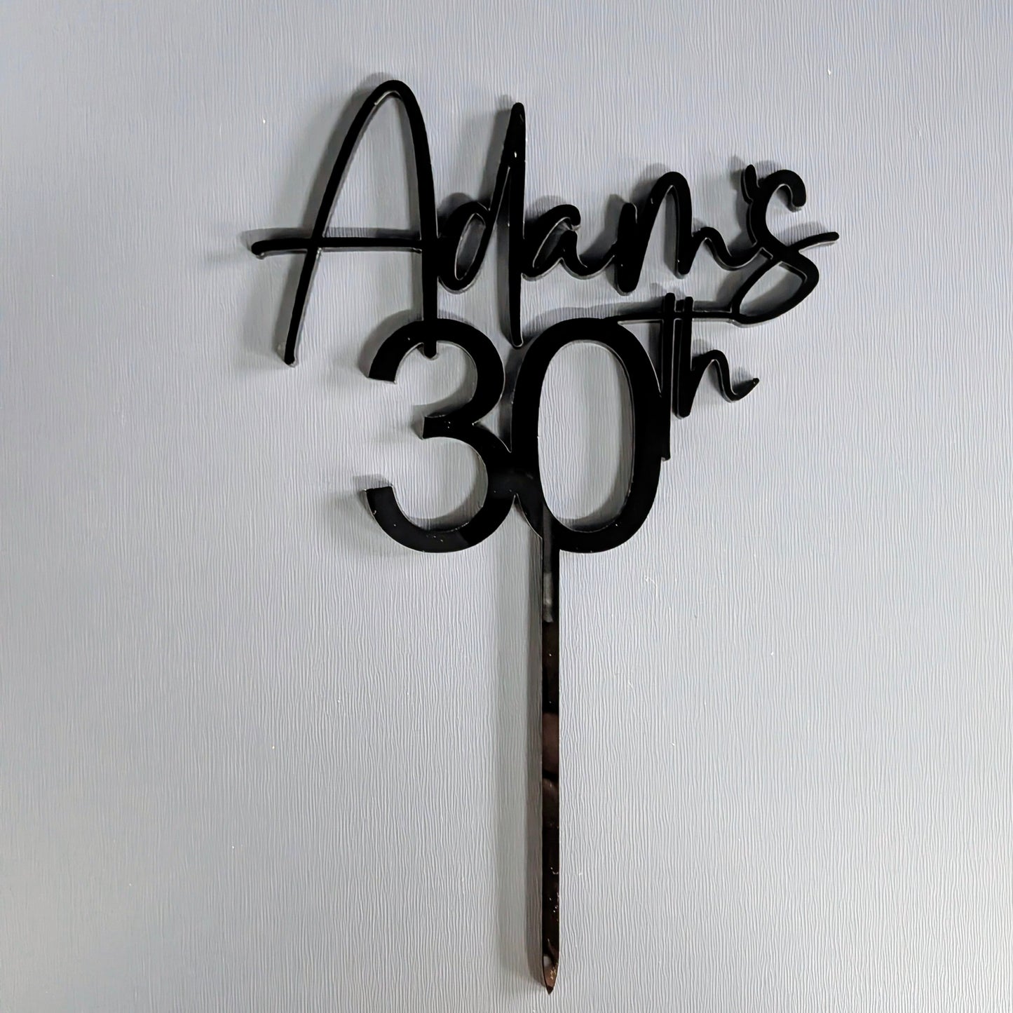 Black cake topper with 'Adam's 30th' on a white background