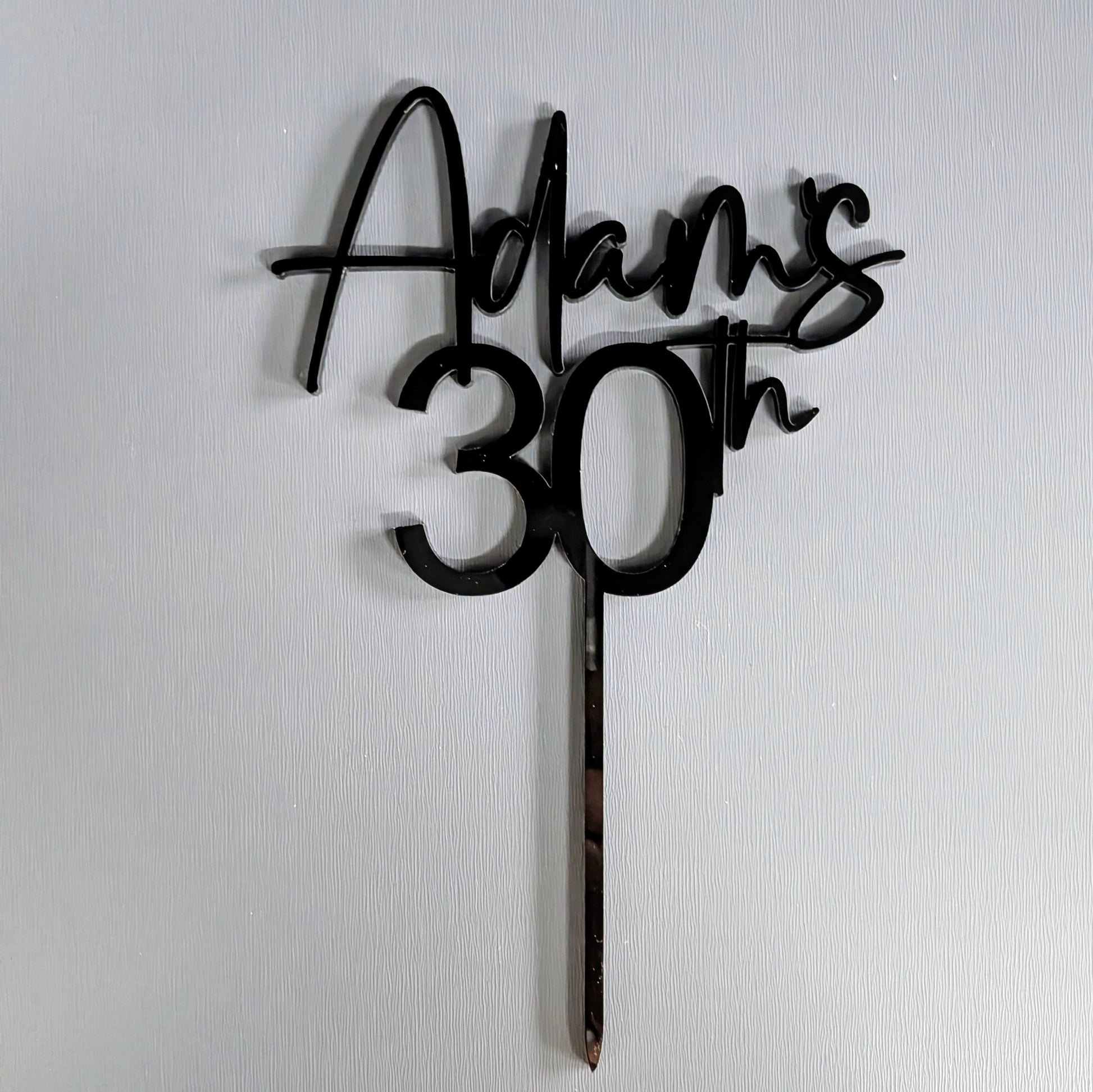 Black cake topper with 'Adam's 30th' on a white background