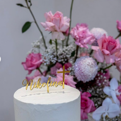 A white frosted cake topped with a gold 'Nikofina' script cake topper and a small gold cross, set in front of a pink rose and floral arrangement against a light gray background.