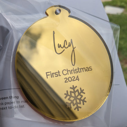 Gold ornament with 'Lucy First Christmas 2024' text on a clear plastic bag.